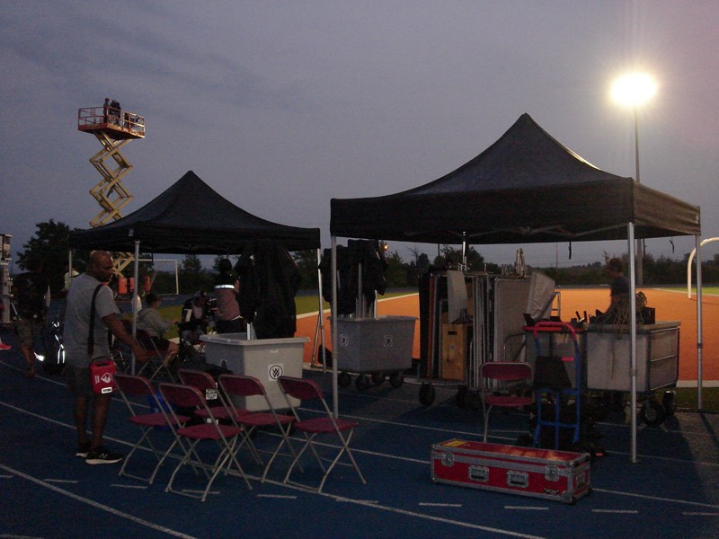 Evening shoot — black pop-up tents, chairs, and equipment carts staged on a sports field with a scissor lift in the background