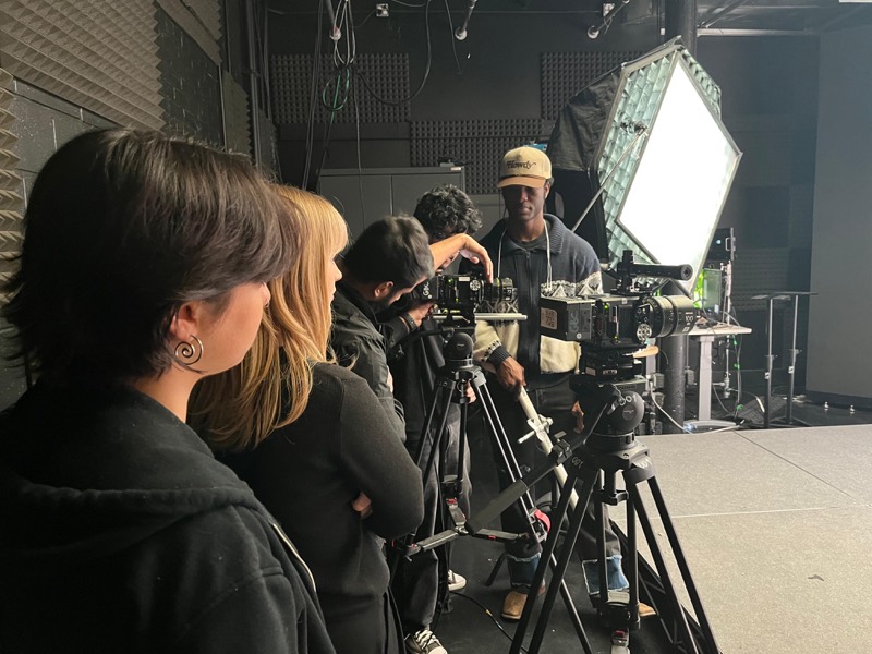 Crew lined up behind cameras on tripods, a large softbox lighting the set
